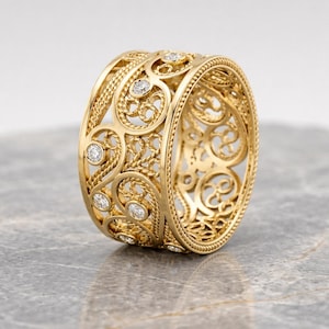 Può includere: Un anello color oro con un intricato design traforato. L'anello presenta una serie di motivi a spirale ed è impreziosito da piccole gemme rotonde e trasparenti. L'anello è esposto su una superficie grigia.