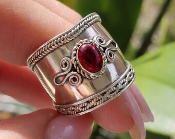 May include: A wide silver ring with an oval, deep red gemstone at the center. The ring features intricate braided and swirl designs. The ring is held up against a blurred green background.