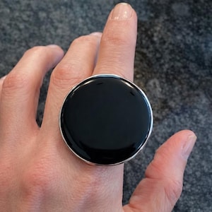 May include: A large, round ring with a glossy black center and a silver-colored border. The ring is worn on a finger, showcasing its bold design. The ring's diameter appears to be approximately 1.5 inches.