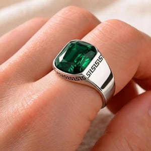 Könnte beinhalten: Ein silberner Ring mit einem großen, smaragdgeschliffenen grünen Edelstein. Der Ring hat ein poliertes Silberband mit einem dekorativen Muster an den Seiten. Der Edelstein hat eine leuchtend grüne Farbe und fängt das Licht ein.