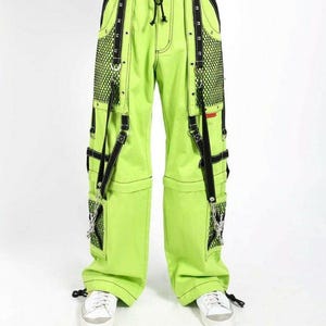 May include: Bright neon green pants with black accents, featuring mesh pockets and decorative straps. The pants have a drawstring waist and are paired with white sneakers. The design includes black stitching and metal hardware.