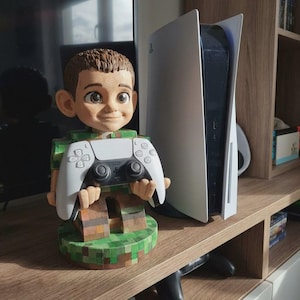 May include: A 3D-printed figurine of a smiling boy holding a white and black game controller. The figurine is on a green and brown base. A white and black game console is next to the figurine on a wooden shelf.