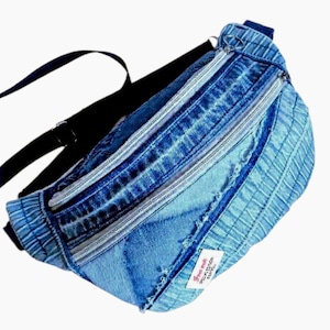 Puede incluir: Un bolso de cintura de mezclilla con múltiples compartimentos con cremallera y una correa negra ajustable. La bolsa está hecha de jeans azules reciclados y presenta una pequeña etiqueta blanca con texto.