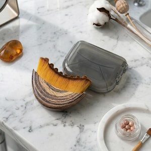 Op de afbeelding: Een make-up kwast met een gebogen, gemarmerde handgreep en een waaier van gele en bruine borstelharen. Een doorzichtige, grijze plastic hoes ligt open naast de kwast. Een klein potje make-up en een kwast zijn ook zichtbaar.