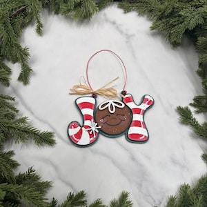 May include: A Christmas ornament spelling "JOY" with a gingerbread man face in the center. The letters are red and white striped, with a small white snowflake. The ornament is trimmed in green and has a raffia bow.