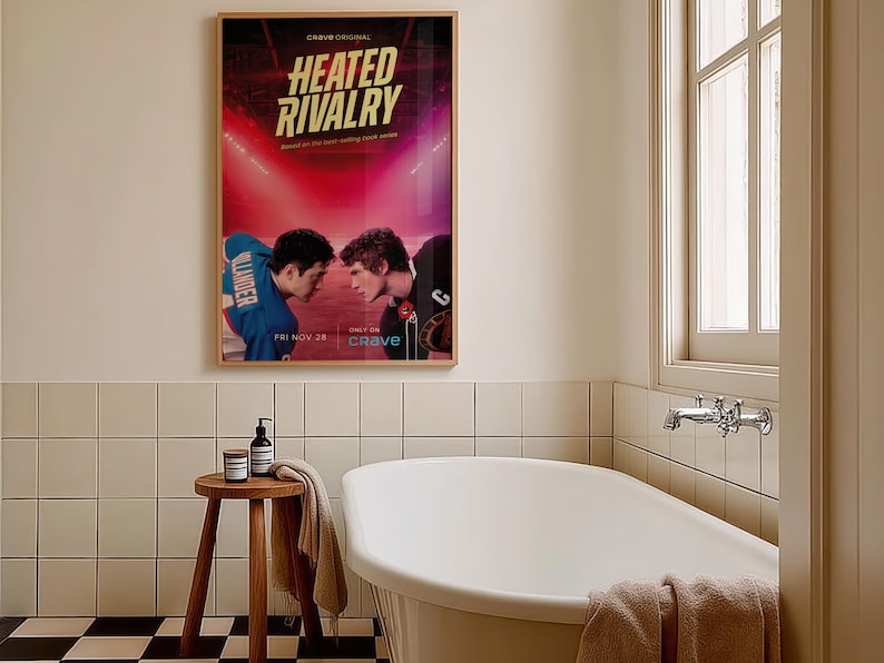 May include: A framed poster for "Heated Rivalry" hangs above a white bathtub in a bathroom. The poster features two football players facing each other. A small wooden stool with a towel and toiletries sits nearby. The bathroom has white tile and a checkered floor.