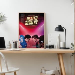 May include: A framed poster for "Heated Rivalry" on a desk. The poster features two men facing each other in a sports arena, with text that reads "Crave Original" and "Fri Nov 28". A laptop, lamp, and camera are also on the desk.