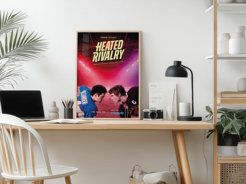 May include: A framed poster for "Heated Rivalry" on a desk. The poster features two men facing each other in a sports arena, with text that reads "Crave Original" and "Fri Nov 28". A laptop, lamp, and camera are also on the desk.