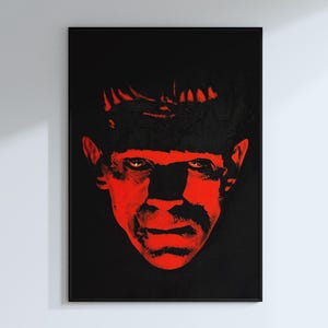 May include: A framed art print featuring a stylized portrait of a monster's face in red against a black background. The artwork has a pop art style, with bold colors and a striking visual impact. The image is mounted in a black frame.