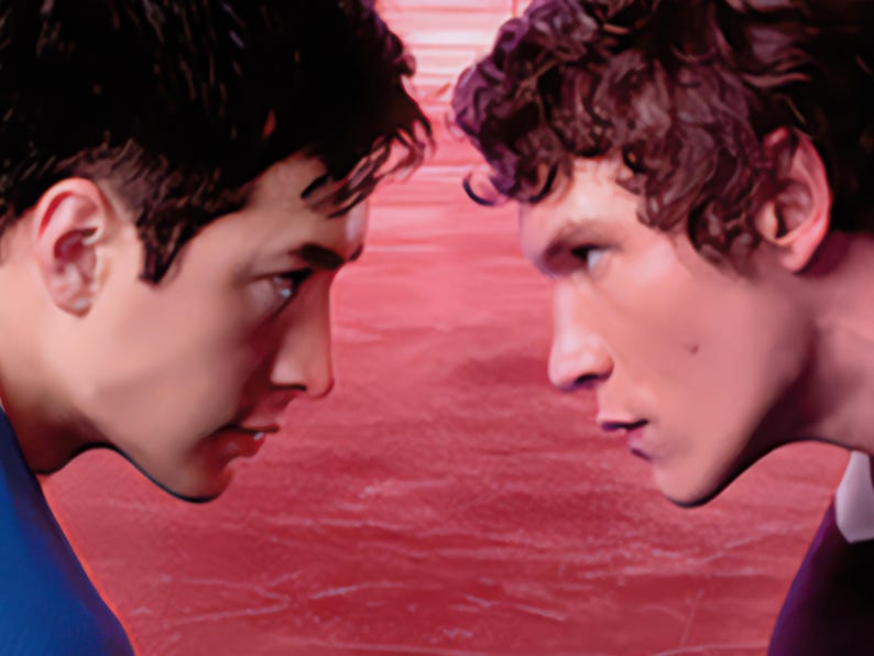 May include: Two young men face each other in a close-up shot, their profiles visible. The background is a textured red surface. One man has dark hair and is wearing a blue shirt. The other has curly hair and a dark shirt.