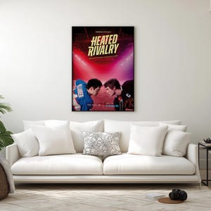 May include: A framed poster with the title "HEATED RIVALRY" in gold lettering, featuring two athletes facing each other. The background is a vibrant mix of red and purple, with bright lights. The poster is displayed above a white sofa.