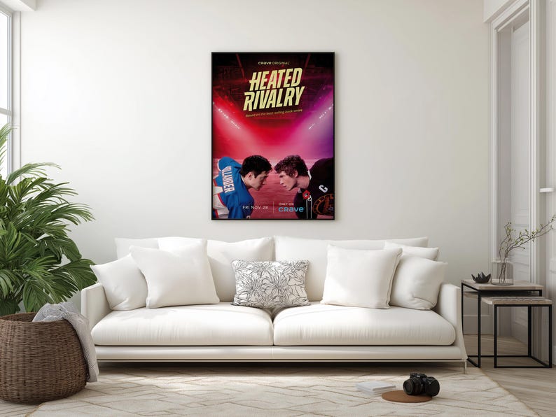 May include: A framed poster with the title "HEATED RIVALRY" in gold lettering, featuring two athletes facing each other. The background is a vibrant mix of red and purple, with bright lights. The poster is displayed above a white sofa.