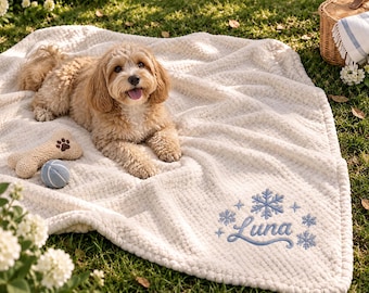 May include: A cream-colored textured blanket featuring the embroidered name Luna in blue script, surrounded by blue snowflake and star designs. A small dog lies on the blanket next to a tan bone-shaped toy and a blue and white ball.
