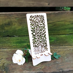 May include: White wooden phone stand with a leafy design cut out. The stand has a small lip on the top to hold a phone in place. The stand is sitting on a wooden surface.