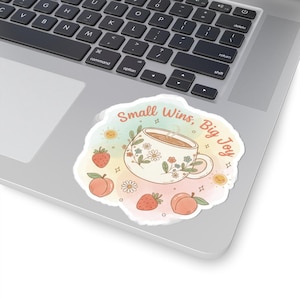 May include: A sticker with a watercolor design featuring a cup of coffee, strawberries, and peaches. The text "Small Wins, Big Joy" is written above the cup. The sticker is placed on a laptop.