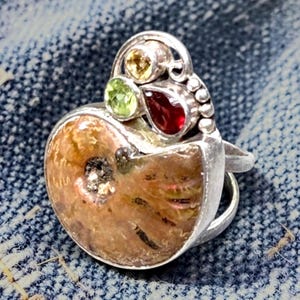 May include: A silver ring featuring a large, circular, brown ammonite fossil as the centerpiece. The setting is adorned with three small, faceted gemstones: a round yellow stone, a round light green stone, and a teardrop-shaped deep red stone.