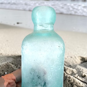 May include: A vintage, translucent light aqua glass bottle with a rounded, bulbous top opening and a cylindrical body. The glass surface shows signs of wear with a weathered, matte texture and small internal air bubbles throughout the material.