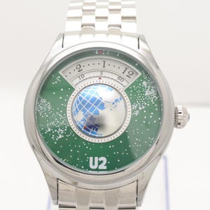 Puede incluir: Un reloj de pulsera plateado con una esfera verde que presenta un diseño de globo terráqueo y el texto "U2". El reloj tiene una correa plateada y una pequeña gema azul en el lateral. La esfera del reloj tiene un diseño circular con números.
