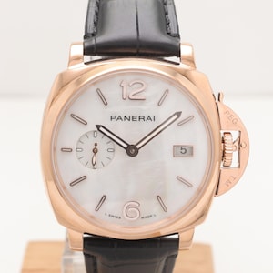 May include: A rose gold Panerai watch with a white face and black leather strap. The watch has a date display at 3 o'clock and a small seconds dial. The watch is displayed on a wooden stand.