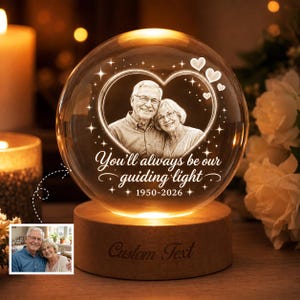 May include: A spherical glass ornament on a wooden base featuring a laser-etched portrait of an older couple inside a heart shape. The text reads, "You'll always be our guiding light" and "1950-2026," with small heart and star accents.