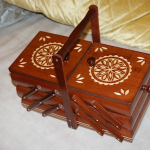 Könnte beinhalten: Eine dunkelbraune Holz-Nähkiste mit einem dekorativen Blumendesign auf dem Deckel. Die Kiste hat einen Griff und ein mehrschichtiges Design mit mehreren Fächern. Sie verfügt über einen Klappmechanismus.