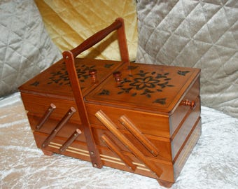 Könnte beinhalten: Eine Holz-Nähkiste mit Griff, mit zweiteiligem Deckel und ausziehbaren Schubladen. Die Kiste ist in einem warmen Braunton gehalten und mit floralen Designs verziert.