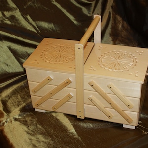 Puede incluir: Una caja de costura de madera clara con asa. La caja tiene un diseño floral tallado en la tapa y soportes de madera en ángulo. La caja está sobre una superficie oscura y texturizada.