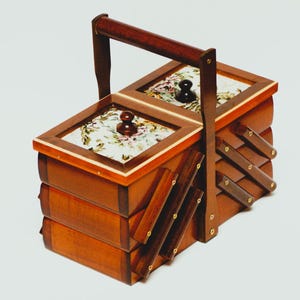 Könnte beinhalten: Eine Holz-Nähkiste mit Griff und zwei Fächern. Die Deckel sind mit floralem Stoff verziert und mit dunkelbraunen Knöpfen versehen. Die Box hat ein gestuftes Design mit ausziehbaren Abschnitten.