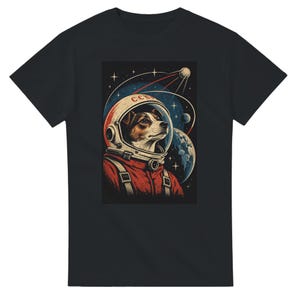 May include: A black t-shirt featuring a graphic print of a dog wearing a space helmet with the text CCCP. The design shows the dog in a red suit against a dark blue space background with stars, a planet, and a satellite orbiting in the distance.