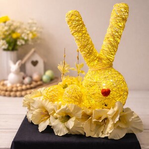 May include: A decorative Easter bunny made of yellow woven material, with illuminated details. The bunny has large ears and a round body, adorned with a red heart-shaped nose. It is surrounded by yellow flowers and sits on a black surface.