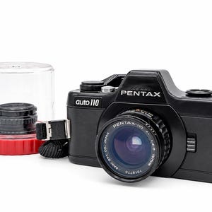 Peut inclure: Un appareil photo argentique noir Pentax auto 110 avec objectif attaché, portant les inscriptions Asahi Pentax et auto 110 sur le boîtier. Un objectif supplémentaire est rangé dans un étui en plastique transparent à base rouge.
