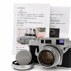 Pode incluir: Uma câmara telémetrica Konica prateada e preta com uma lente Hexanon de 50mm f/1.8, uma tampa de lente circular de metal com o logótipo Konica, um para-sol prateado e um manual de instruções branco com texto em japonês e diagramas.