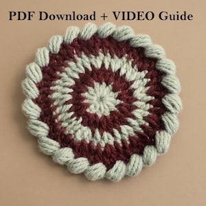 May include: A handmade, circular crochet coaster with a spiral design. The coaster features a central pattern of burgundy and cream-colored yarn, surrounded by a border of light green yarn. The text "PDF Download + VIDEO Guide" is at the top.