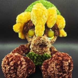 May include: A handmade plush toy with a whimsical design. The toy features a brown body, yellow and green accents, and large, yellow-green eyes. The plush has a soft, textured appearance, suggesting a cozy feel.
