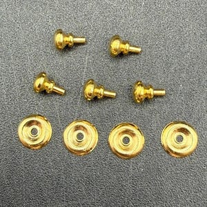 May include: A collection of nine gold-colored metal hardware components consisting of five small studs with rounded heads and protruding stems, alongside four flat, circular washers with a central hole, all arranged on a textured surface.