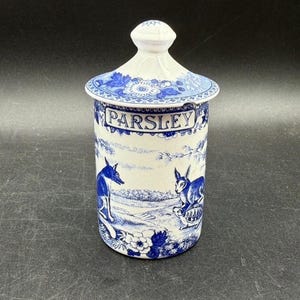 May include: A white and blue ceramic parsley jar with a lid. The jar features a blue and white pastoral scene with animals and the word "PARSLEY" in a banner. The lid has a floral design.