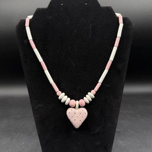 May include: A pink and white beaded necklace with a heart pendant. The necklace features alternating white and pink cylindrical beads, with a heart-shaped pendant adorned with black polka dots. The necklace is displayed on a black stand.
