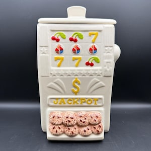 May include: A cream-colored ceramic cookie jar shaped like a vintage slot machine. The front features raised relief details including red cherries, yellow sevens, and blue flame icons. A yellow dollar sign sits above the word JACKPOT in yellow letters.
