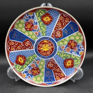 May include: A decorative ceramic plate with a vibrant floral design. The plate features a circular pattern with red, blue, green, and yellow segments. The center has a yellow flower. The plate is supported by a clear plastic stand.