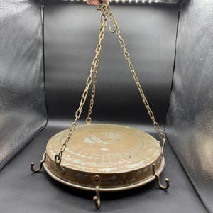 May include: A vintage copper cooking pot with a round, flat base and a hanging chain. The pot has three curved hooks around the rim and is suspended from a chain with a hook at the top. The copper has a weathered, aged appearance.