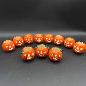 May include: A collection of twelve small orange ceramic jack-o'-lantern candle holders. Each piece features a carved face with a glossy finish. Three of the pumpkins in the front row have small green painted leaves attached to the top rim.