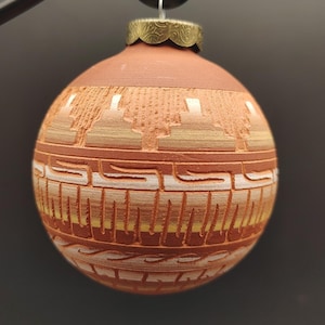May include: A spherical terracotta ornament featuring carved horizontal bands with geometric patterns in white and yellow. The ornament has a metallic gold-toned cap at the top with an embossed swirl design and a silver hanging loop.