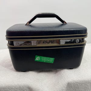 May include: A black Samsonite hard-shell vanity case with a textured finish, a top handle, and two silver-toned metal latches with key locks. A green rectangular security sticker is adhered to the front, featuring the text SECURITY CHECKED.