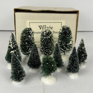 May include: A set of miniature frosted green Christmas trees with white detailing. The trees, of different sizes and shapes, are on white bases. The box behind the trees reads "Village Frosted Trees".