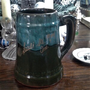May include: A green and brown ceramic beer stein with a handle. The stein has gold lettering that reads "Sauli Ste. Marie Ont."