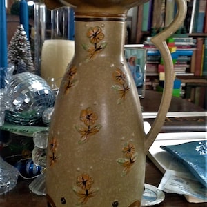 May include: A brown ceramic jug with a wide mouth and a handle. The jug is decorated with a floral pattern in shades of brown and yellow. The jug has a scalloped rim and a brown and white striped base.