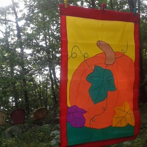 May include: A colourful autumn-themed fabric banner featuring a large orange pumpkin with a green leaf and a yellow stem. The pumpkin is surrounded by smaller green, purple and yellow leaves. The banner has a red border and a yellow background.