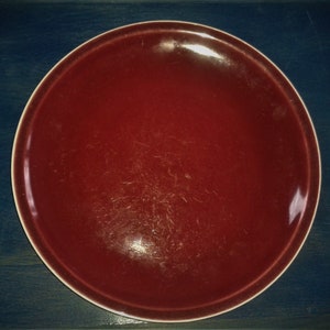 May include: A deep red ceramic plate with a slightly glossy finish. The plate has a white rim and shows signs of wear.