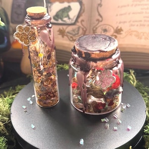 May include: A tall, thin glass vial and a short, wide glass jar, both filled with assorted colorful beads and charms, topped with cork stoppers and brown wax drips. Each container features a small gold-toned paw print charm attached to the neck.