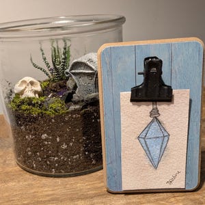 May include: A glass terrarium with a miniature skull and a small house, next to a wooden clipboard with a blue background and a drawing of a blue crystal. The clipboard has a black clip at the top.
